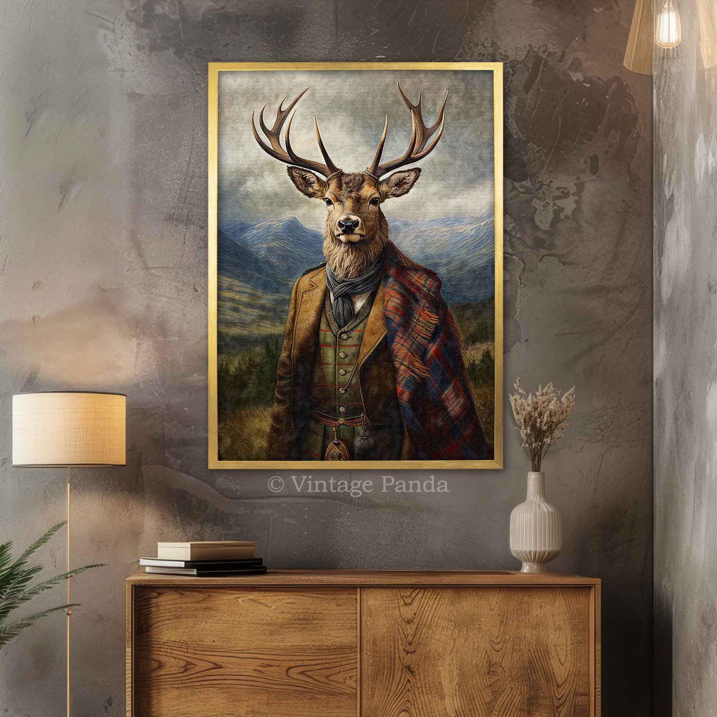 Framed artwork of a deer wearing a coat with Scottish highland mountains in the background, displayed on a wall above a wooden cabinet in a gold frame.